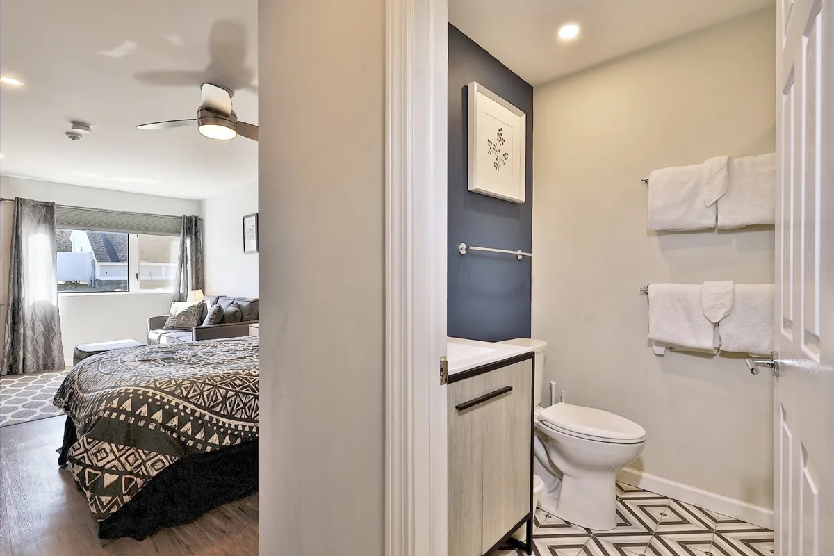 This image showcases a cozy studio interior featuring a bed with patterned bedding, modern bathroom vanities, and decorative wall art. Natural light fills the space, and the layout hints at an all-in-one room ideal for both relaxation and productivity.