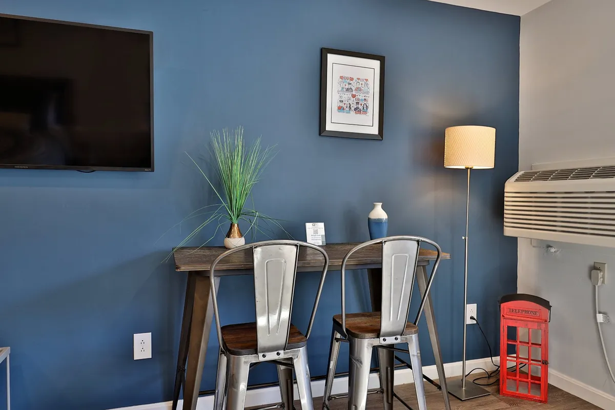 This image displays a modern, relaxed dining area featuring a wooden table with metal chairs, surrounded by framed artwork, a table lamp, a green plant, and a playful red telephone booth accent. The space is bright and conducive to casual dining, working on a laptop, or socializing, echoing the section's mention of multifunctional meal moments.