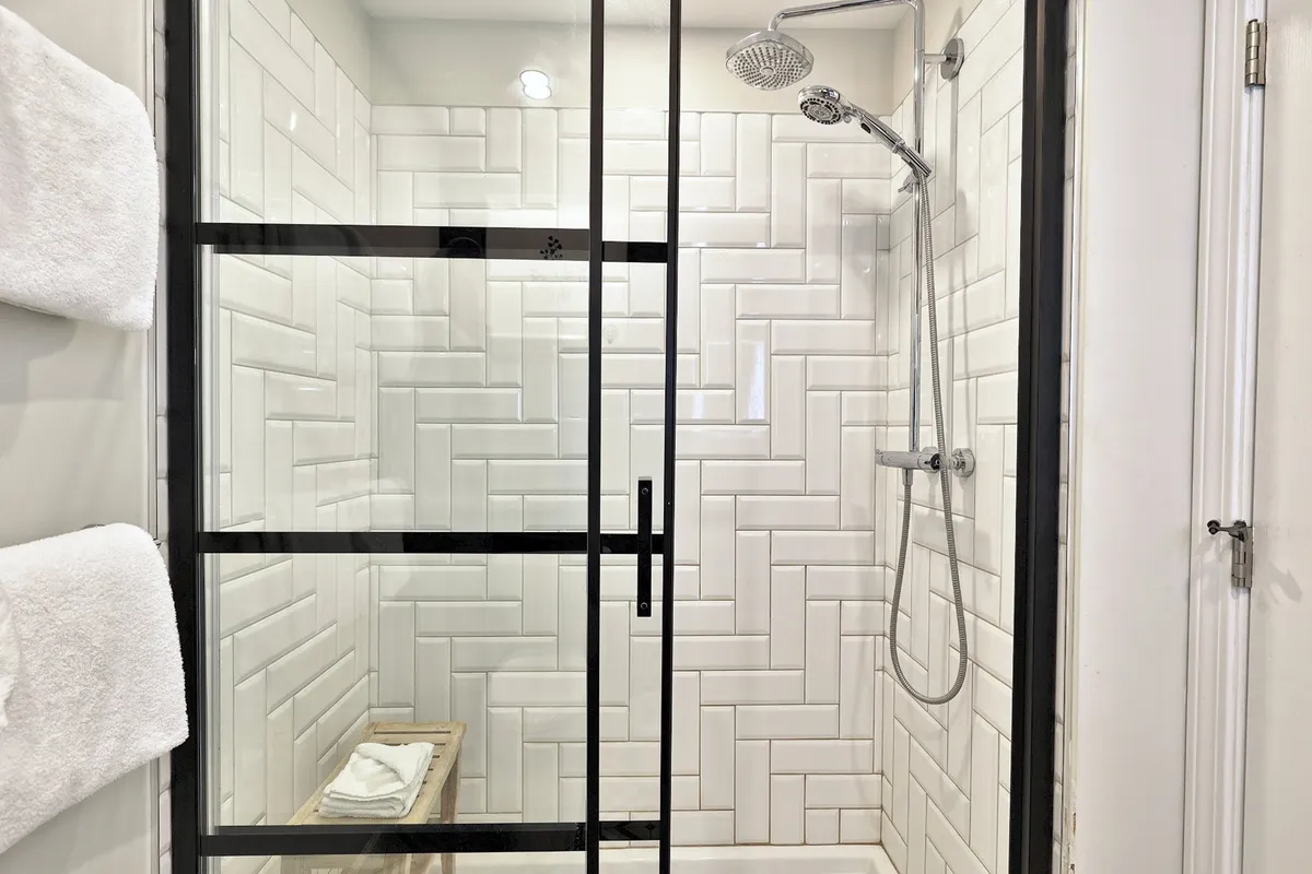 The image highlights a spa-like ensuite bathroom with a shower featuring black accents and a herringbone tile pattern. A wooden stool stocked with towels completes the scene, evoking a sense of rejuvenation and relaxation.