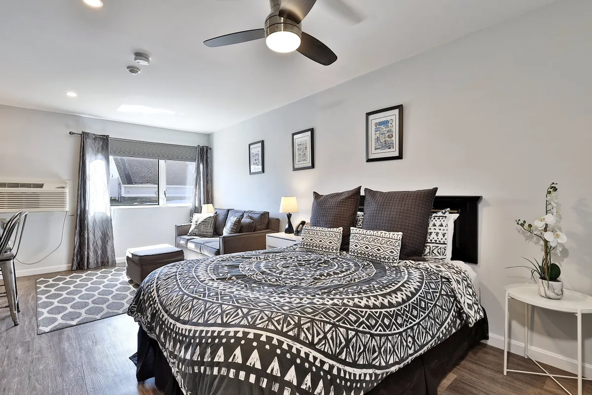 A decorative bedspread adorns the comfortable bed, accompanied by a sofa and a seating area. Artwork decorates the walls, and generous natural light streams in through the window, creating a peaceful, cozy atmosphere ideal for unwinding or socializing.