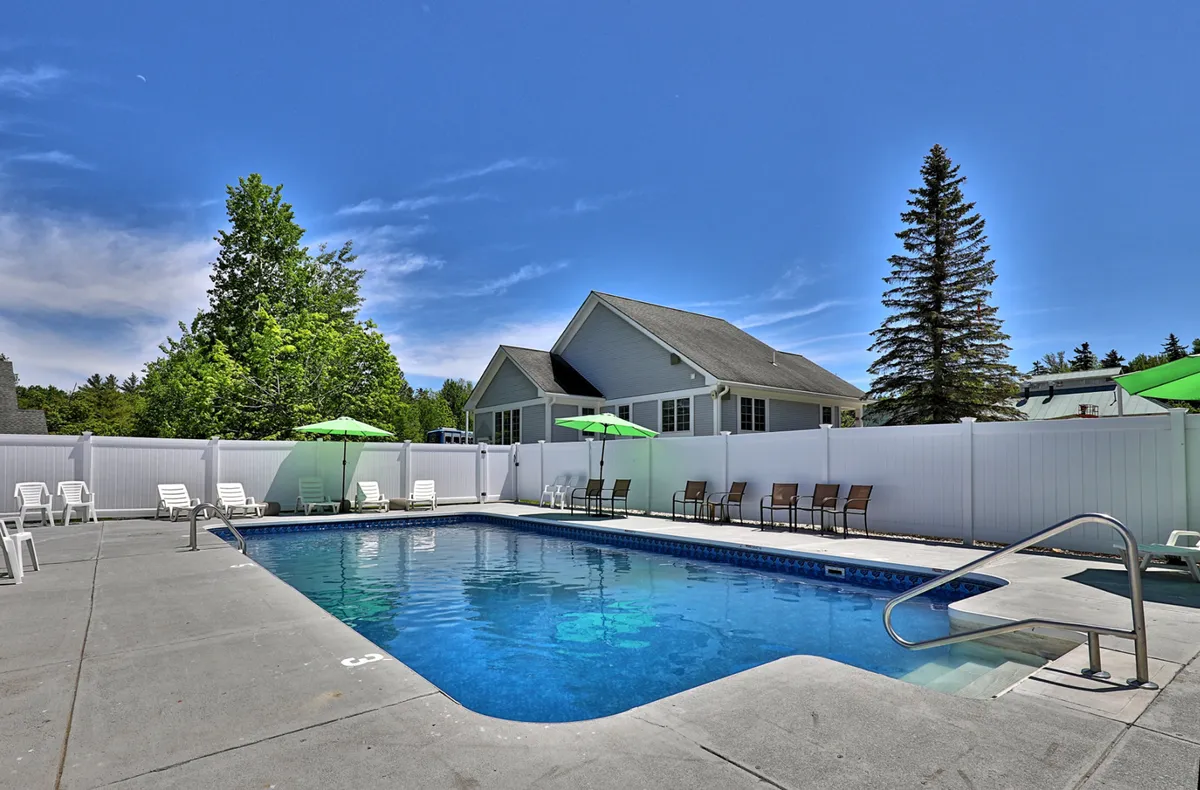 This exterior scene features a sparkling swimming pool surrounded by green umbrellas, lounge chairs, and a crisp white fence. The pool area is framed by lush trees, giving it a relaxing and resort-like ambiance. The setup invites visitors to swim, sunbathe, or gather with friends for a leisurely afternoon under the sun.