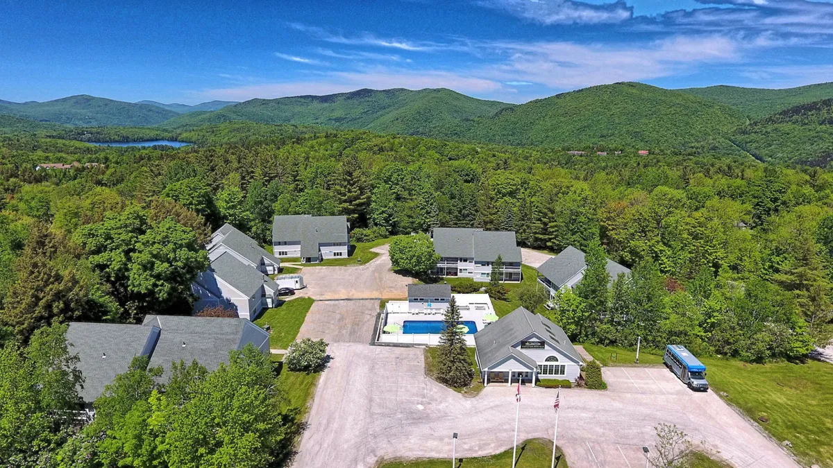 This exterior image showcases the main building of the resort-style complex, featuring a pool area and the surrounding mountains in the background. The parking area is also visible, reinforcing the property's proximity to outdoor amenities and mountain activities.