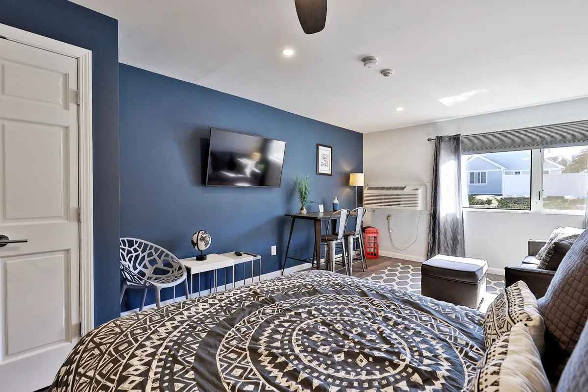 This image features a plush queen bed with patterned bedding, a wall-mounted smart TV, a small dining table with chairs, and a window allowing natural light to fill the room. The overall mood is inviting and comfortable, with touches that reflect recent renovations and modern décor.