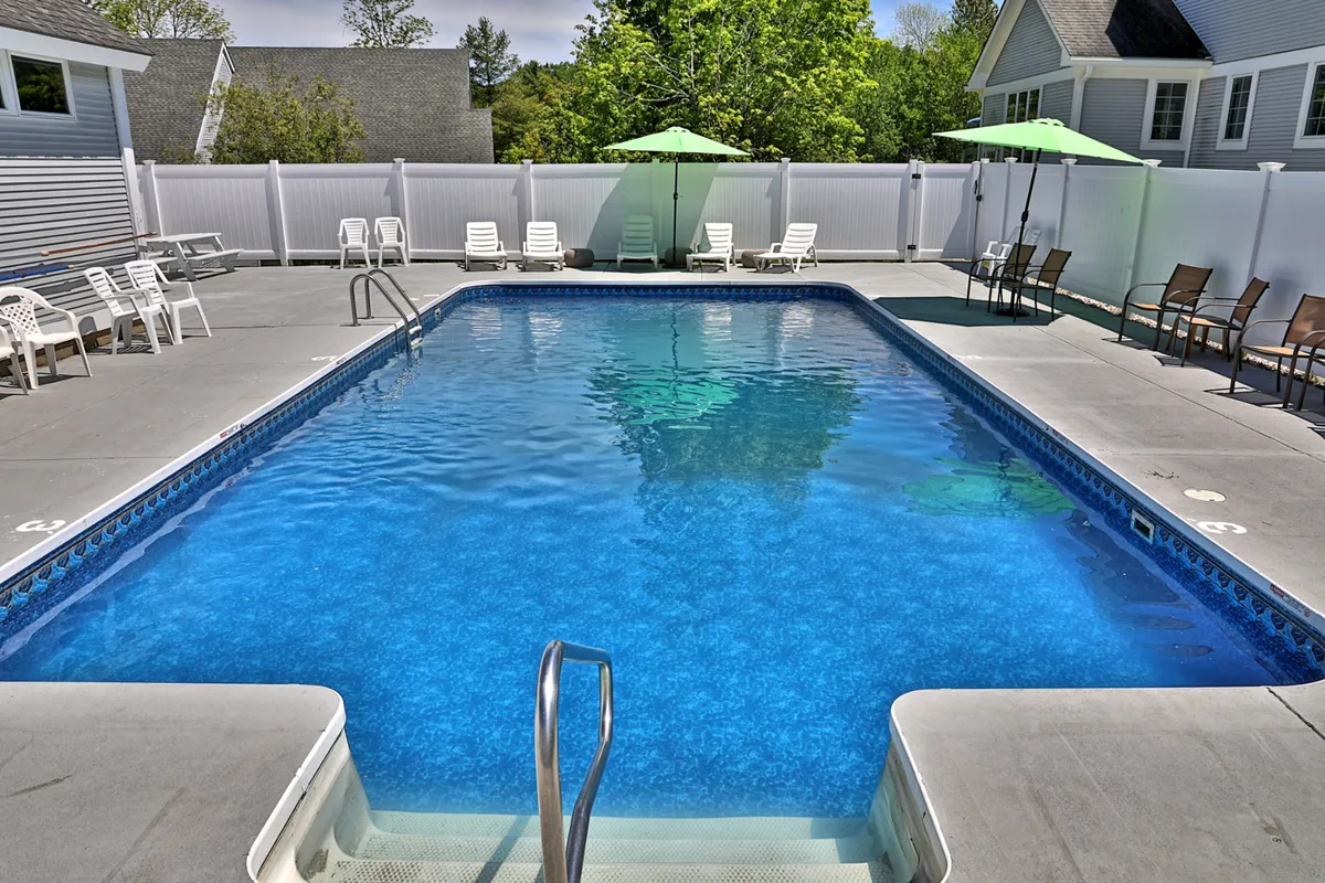 A sunlit outdoor swimming pool surrounded by lounge chairs and umbrellas, bordered by lush greenery. The inviting pool area suggests a space for summer relaxation and socializing just steps away from the condos.
