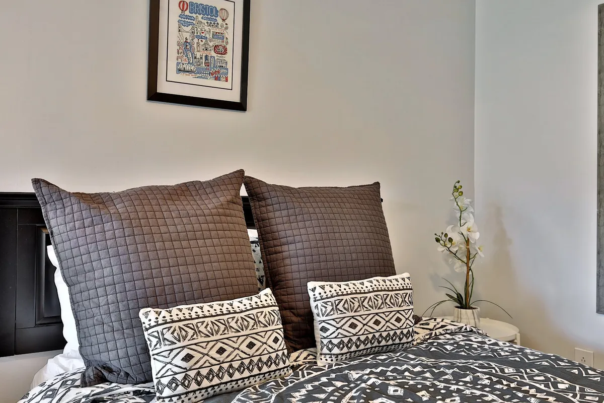 A close-up of a bed with large quilted pillows and decorative cushions, complemented by framed wall art and a potted plant. The scene radiates comfort and a welcoming vibe, ideal for unwinding or enjoying quiet moments.