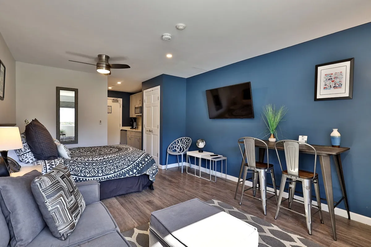 The image showcases a stylish, renovated studio interior: a plush queen bed with patterned bedding sits beside a modern sleeper sofa adorned with decorative pillows. A compact dining table with stools is perfectly placed for meals or work, while a wall-mounted TV invites cozy movie nights. A touch of greenery on the table and sunlight by the window enhance the welcoming, relaxed vibe.